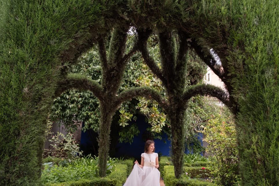 Fotografía de una niña de comunión bajo unos setos de un jardín en el exterior
