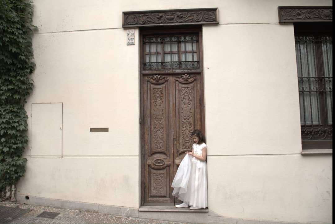 Fotografía de una niña de comunión de pie apoyada en un portón en una casa de Granada